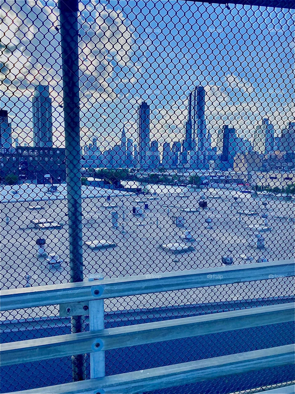 Looking at the “Manhattan” skyline and partly also the silhouettes of “LIC, Queens” from the “Pulaski Bridge” at “Newtown Creek” from the “Greenpoint, Bklyn” side thru a mesh metal fence over a large flat rooftop. 2022. Hypnotic Productions 