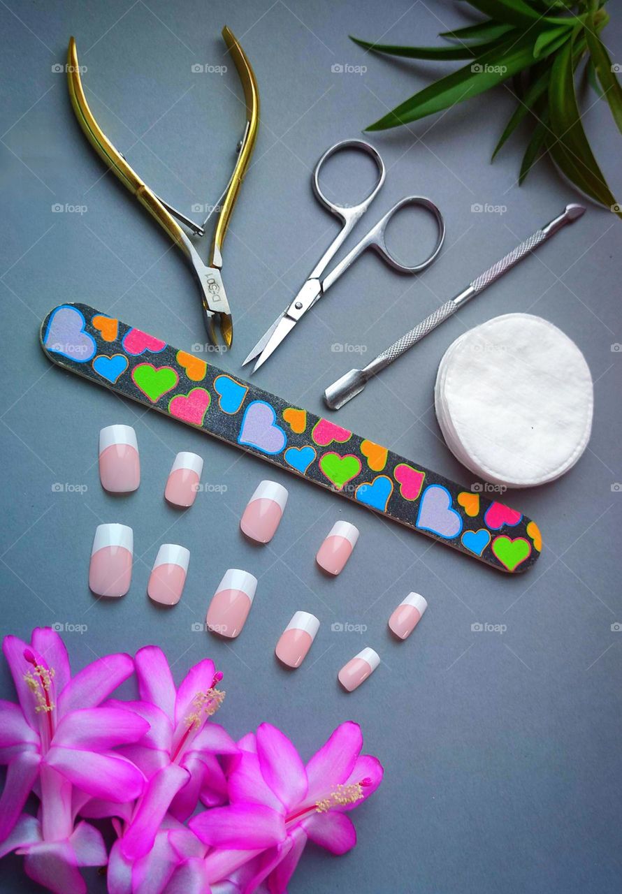 Manicure.  The gray background is divided by a multicolored nail file.  On one side are false nails and three pink cactus flowers, on the other side are: metal nail scissors, cuticle nippers, spatula, cotton pads and green leaves
