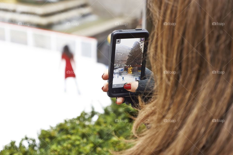 woman taking picture with smartphone of ice skater at rockefeller center