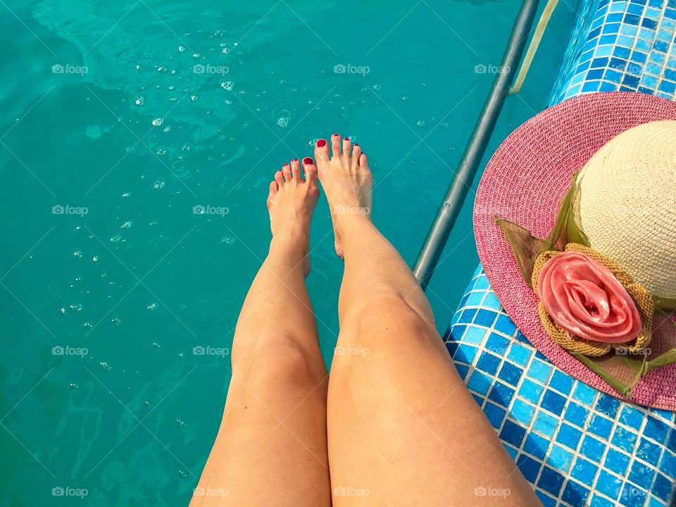 Woman's legs in the swimming pool with pink summer hat beside