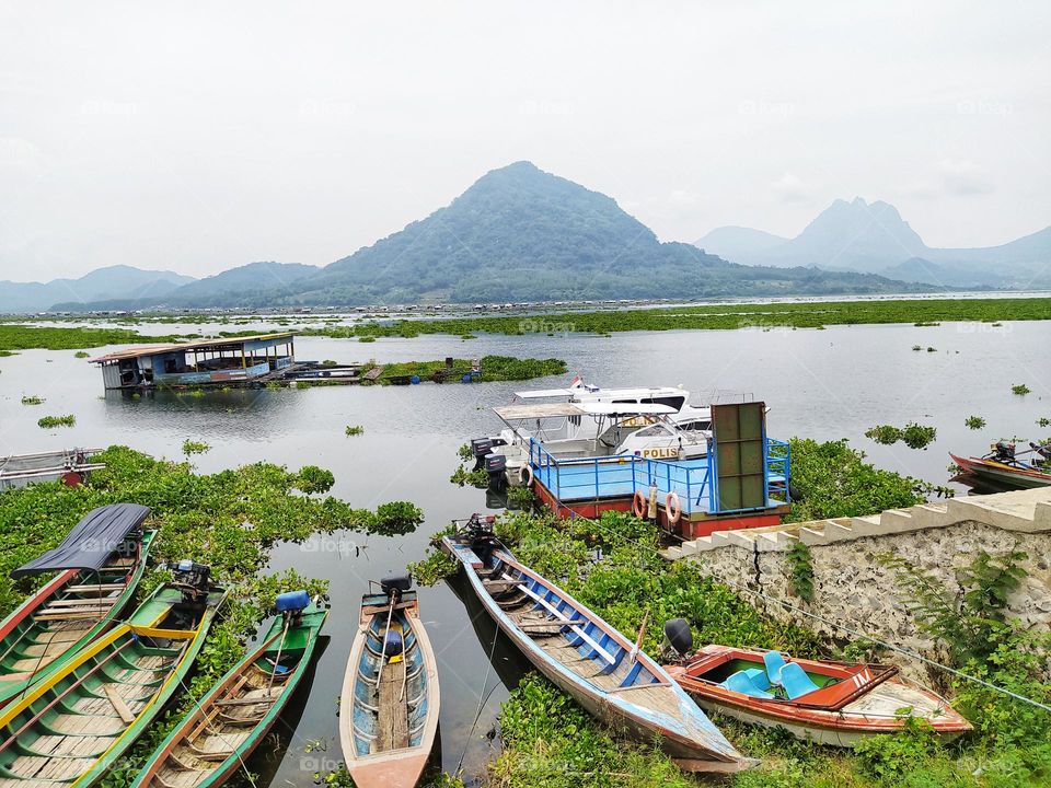 A very beautiful lake view with a mountain background, water hyacinth plants on the surface of the lake and fish farmer boats on the shore