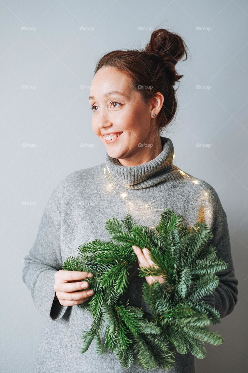 Adult beautiful smiling woman forty years with brunette curly hair in warm grey knitted dress with diy fir christmas wreath in hand on grey background