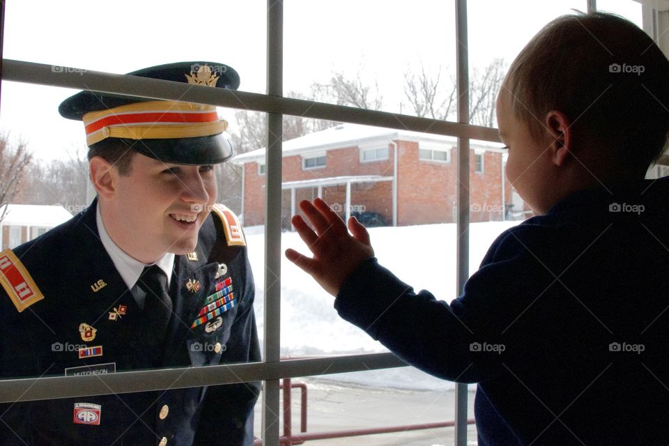 Father in military uniform looking in window as toddler son looks out 