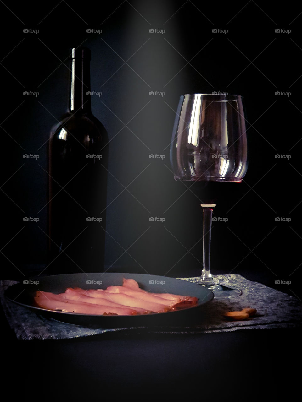 Dark still life.  Black bottle of wine, a glass with red wine and a black plate with dried meat on a decorative ornamental napkin on a black background