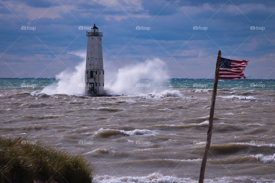An American Flag blowing in the wind, next to a lighthouse on a very windy day.