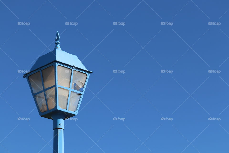 Vivid clear blue sky is the vivid backdrop for this vintage blue street lamp lantern with broken glass inserts likely vandalised by flying rocks 