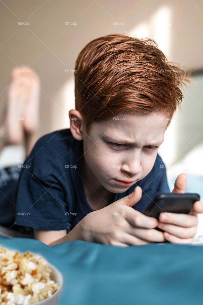 Red-haired boy watching a movie on the phone lies on the bed