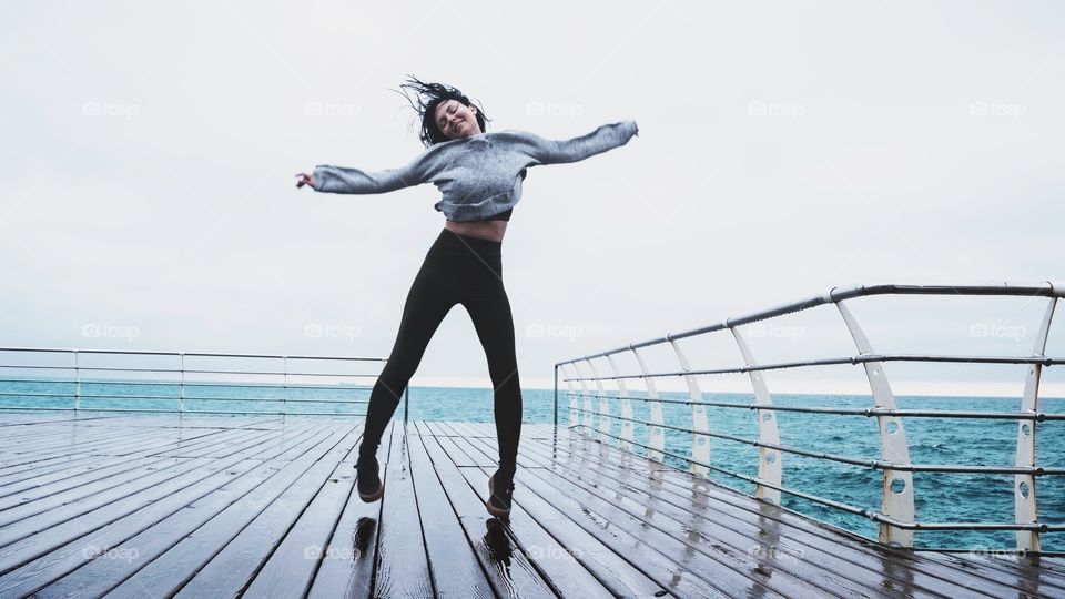 Beautiful slim woman in sports clothing dancing on pier seaside under rain