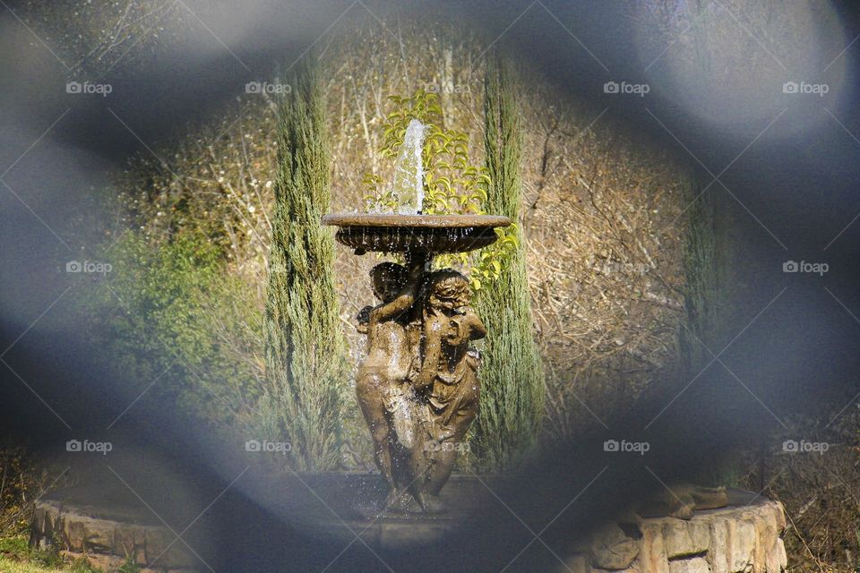 Fountain framed through a patterned chair. Focus your attention on the beautiful fountain held up by the two angels