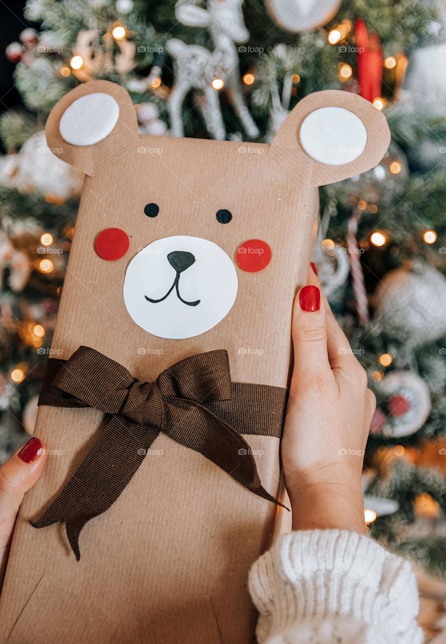 Close-up image of woman's hands holding wrapped christmas present shaped as a teddy bear in front of christmas tree