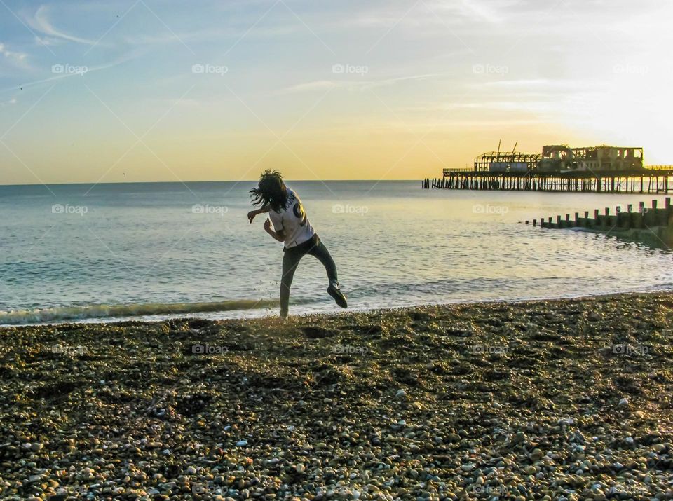 A young man in a football shirts leaps on the beach as he throws stones into the sea in the late afternoon sun, near Hastings pier