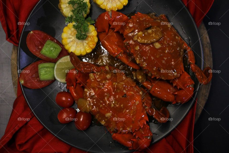 Crab flavored in creamy garlic sauce served with corn, tomatoes, lemon wedges and cherries on a black plate set on a red cloth set in a wooden setting. Asian flavor crab.