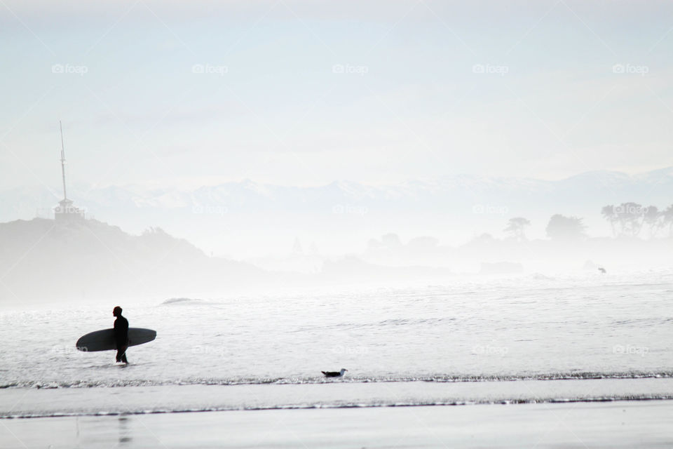 Silhouette of a surfer walking with surfboard on foggy sea 