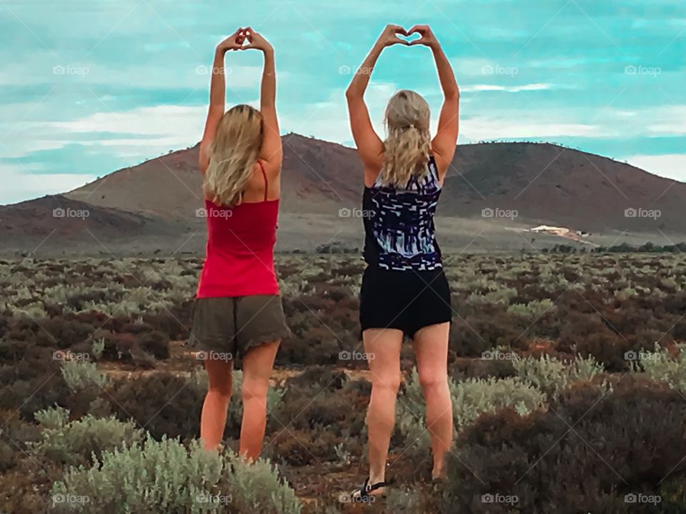 Two blonde young women in the Australian outback, heart shaped with hands, facing sky and mountain