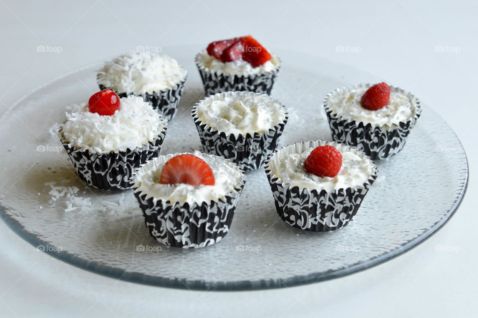 Mini cheesecakes in black and white decorative wrappers topped with whipped cream, coconut shavings, and berries