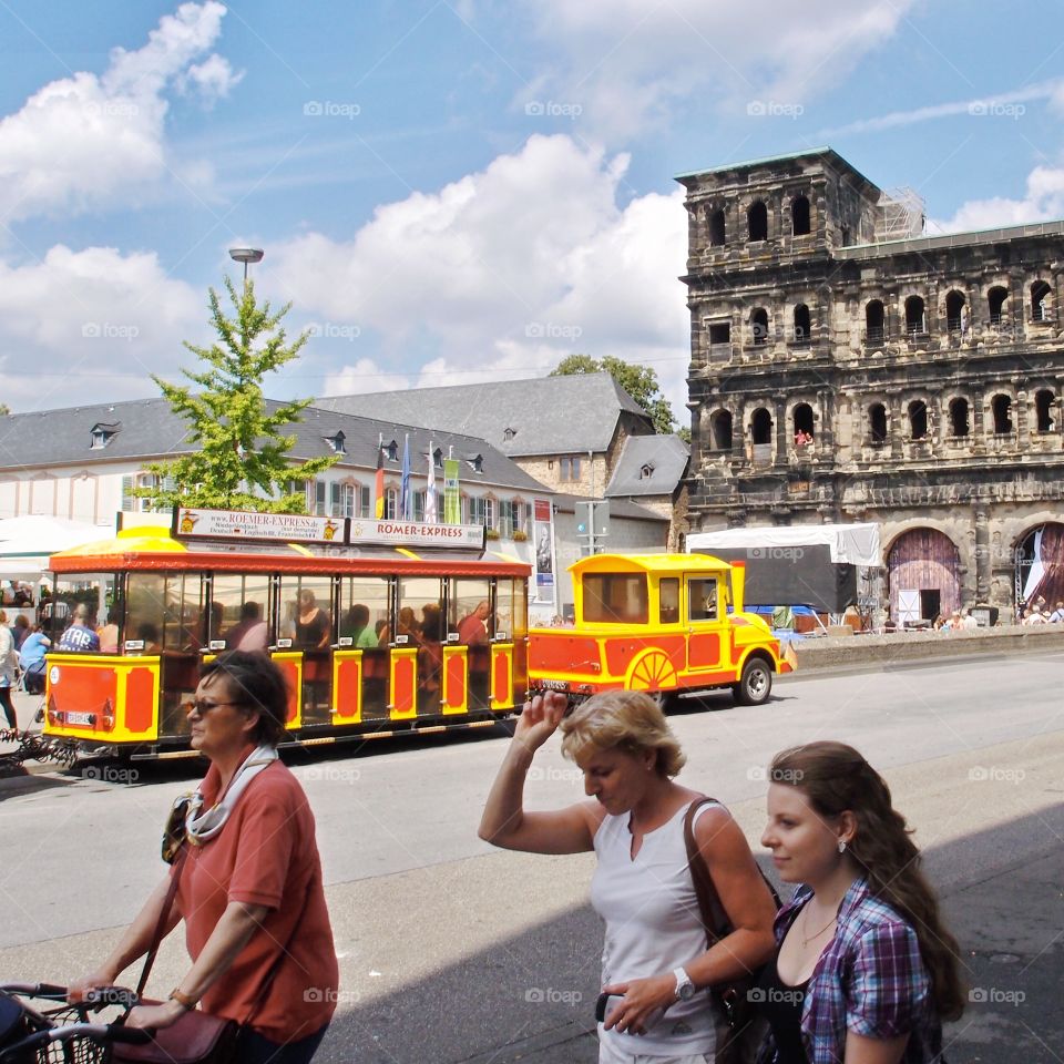 A train style transport runs through a street with walking people and an old stone building in the background on a nice summer day in Europe. 