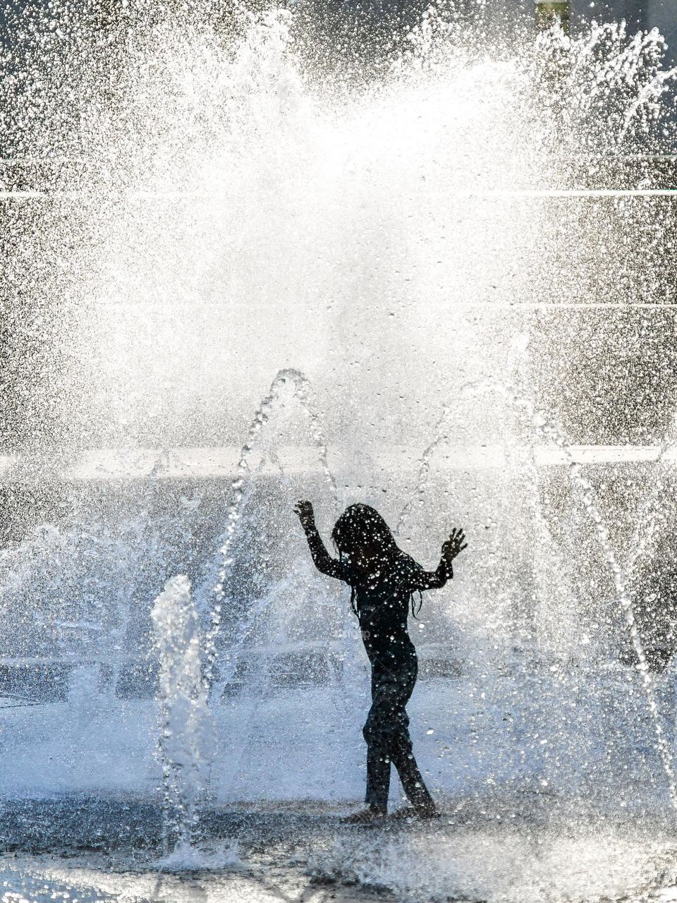 A girl silhouette playing in the water fountain against a bright sunny hot day
