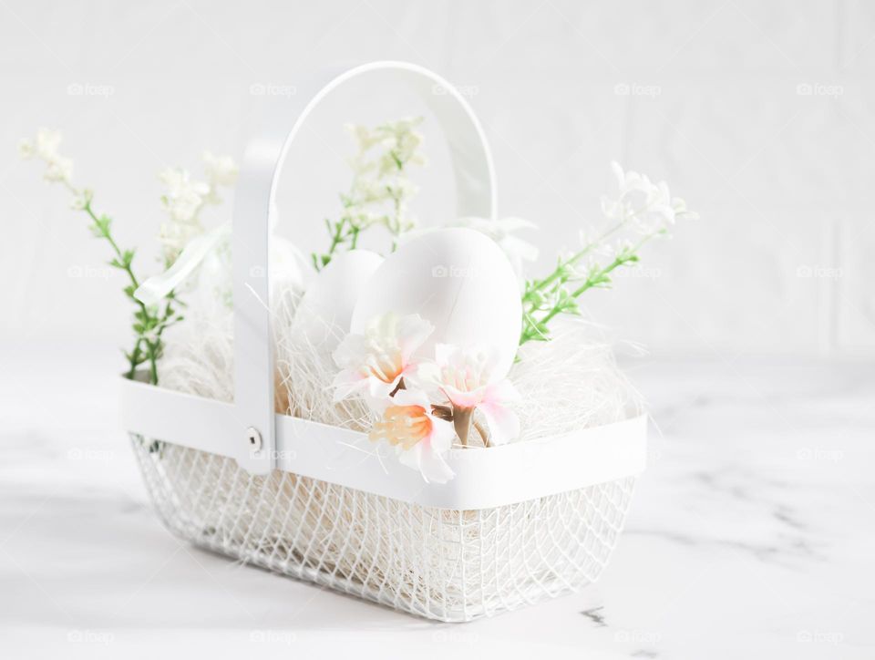 A white wicker metal basket with eggs and twigs with flowers stands on a marble table, side view, close-up.