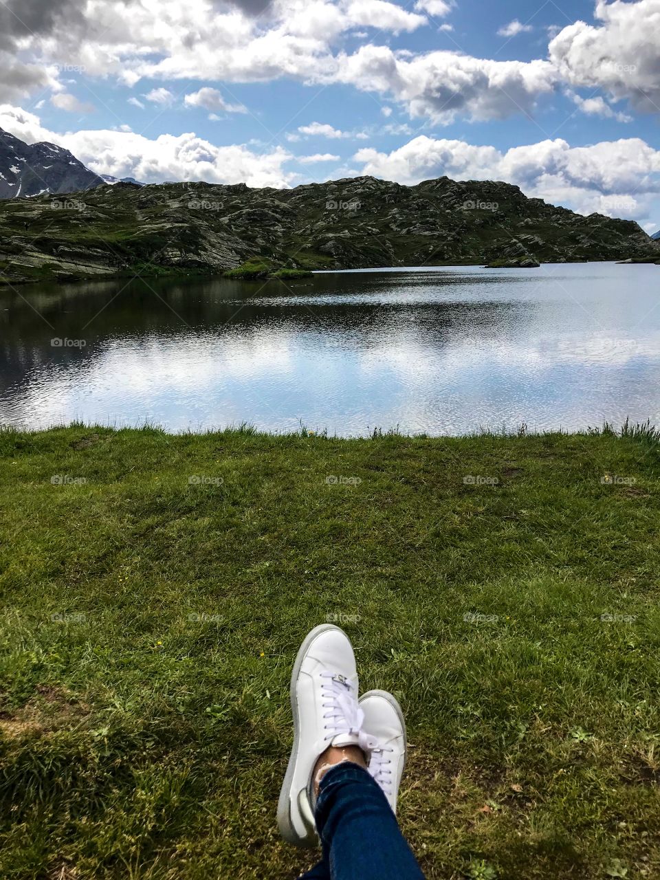 I sitting on the green grass, watching the natural landscape, mountains, lake, in the blue sky have many white clouds. Its shadows are miraculously reflected in the water, and my feet with blue jeans and white shoes