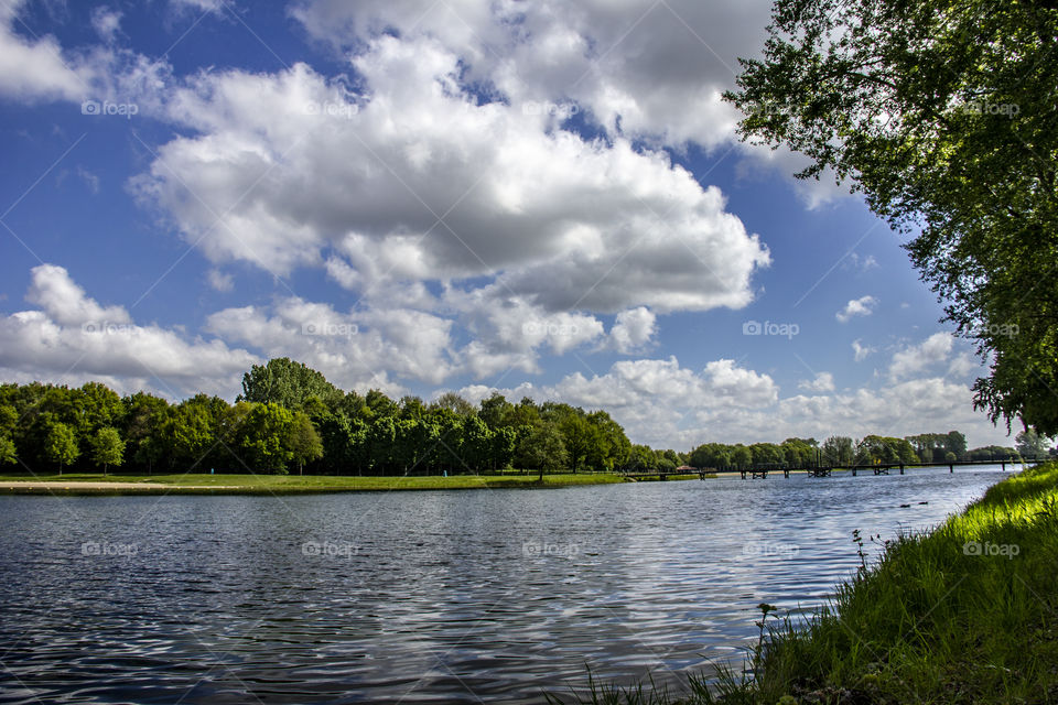 A landscape photo of the lake in bussloo in Holland. the clouds reflect in the riply water of the lake. the lake is surrounded by a forest. there is also a bridge to easily cross the lake.