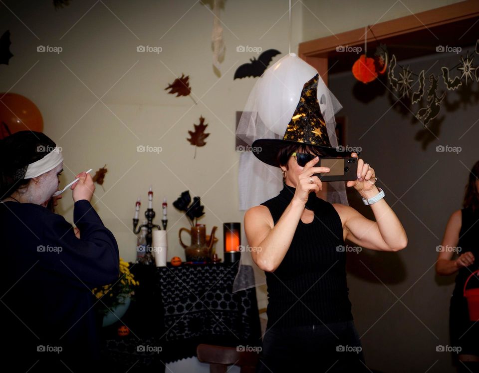 Halloween party.  A girl dressed as a witch takes pictures of the party on the phone.