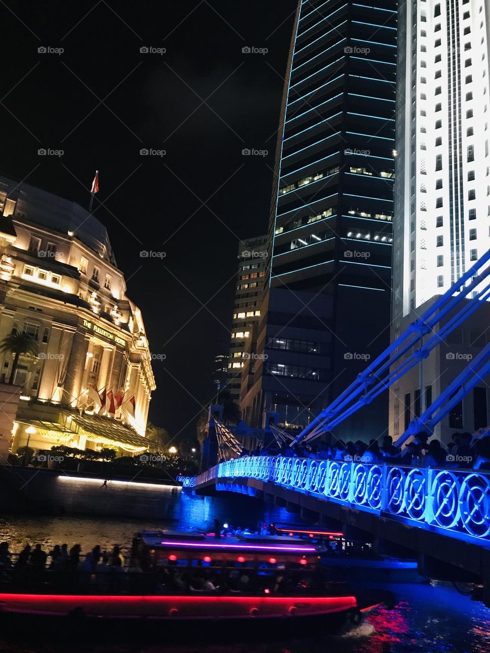 The bridge on river with beautiful night lightings and tourist boat under the bridge along with tourists near the Fullerton hotel in Singapore
