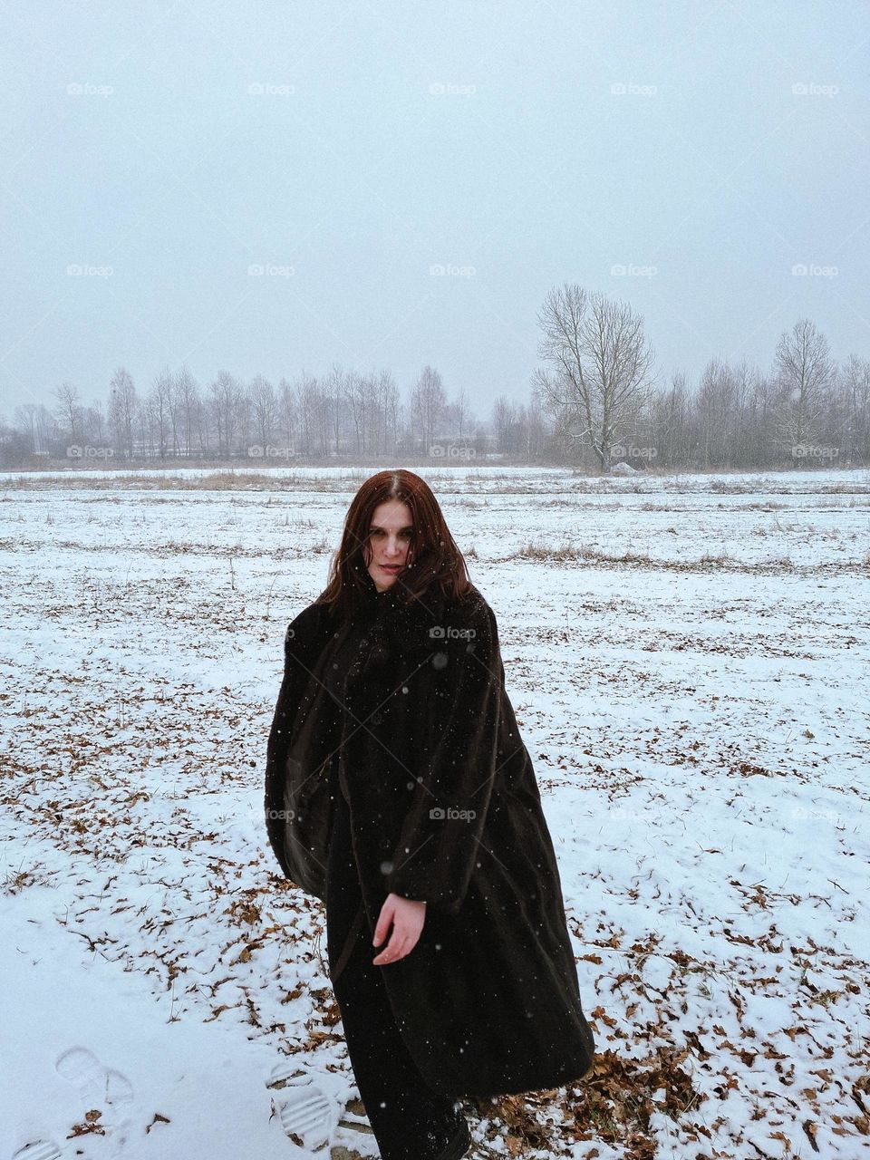light-skinned girl in a dark fur coat stands in a snowy field during a snowfall and waves her fur coat.