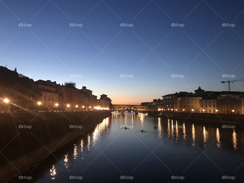 Row boats sailing along the Arno river under the Ponte Vecchio lit by the city lights and the evening moon.