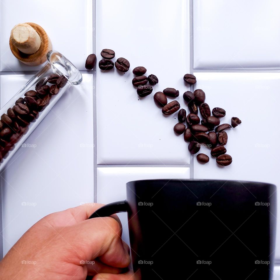 Coffee beans spilling out of slender glass container into a black coffee mug being held by a hand.  This is on a vertical white kitchen tile background.