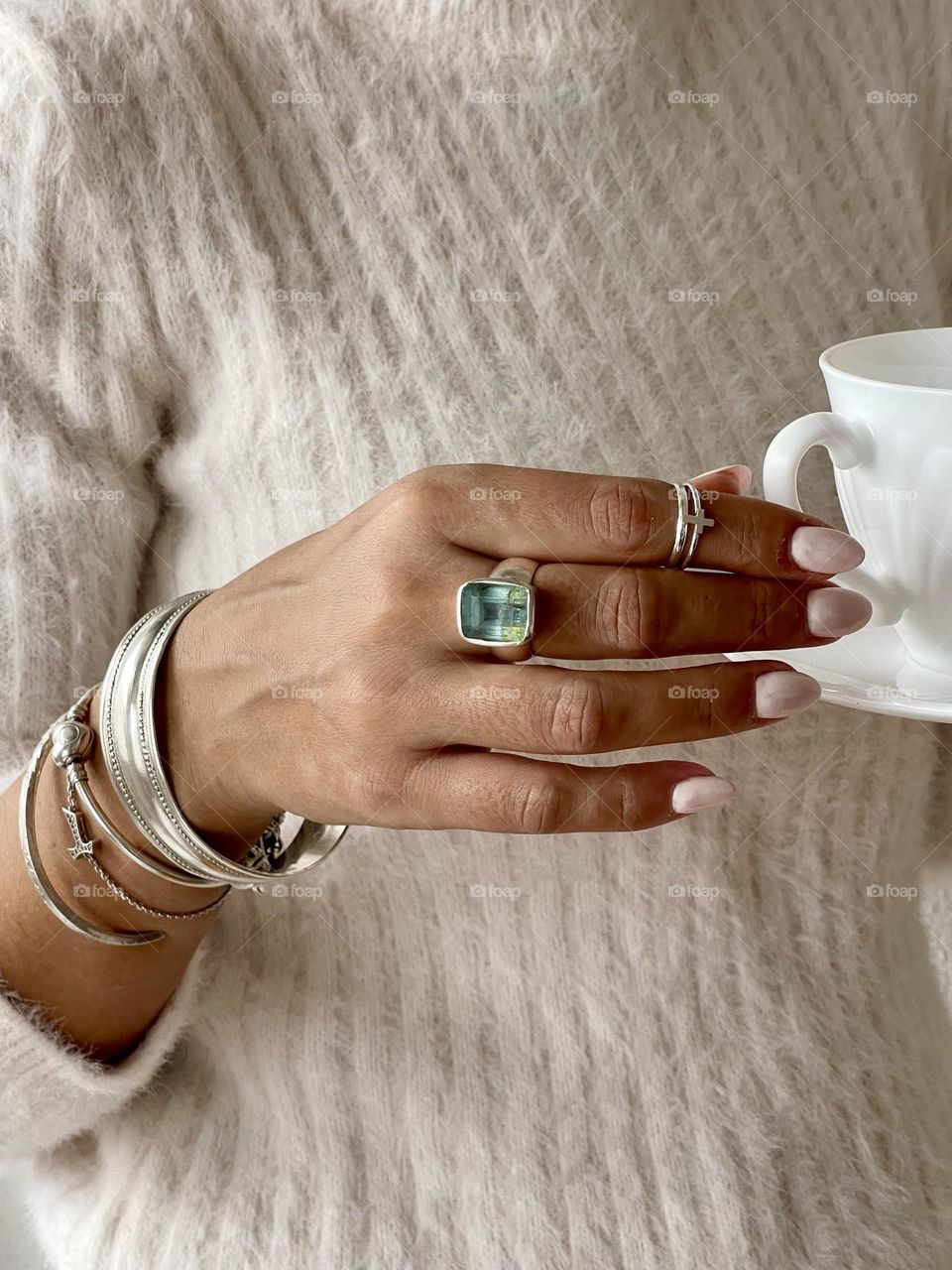 Woman’s hand wearing white nail polish manicure, silver rings with aquamarine stone, silver bracelets holding a white ceramic coffee cup 