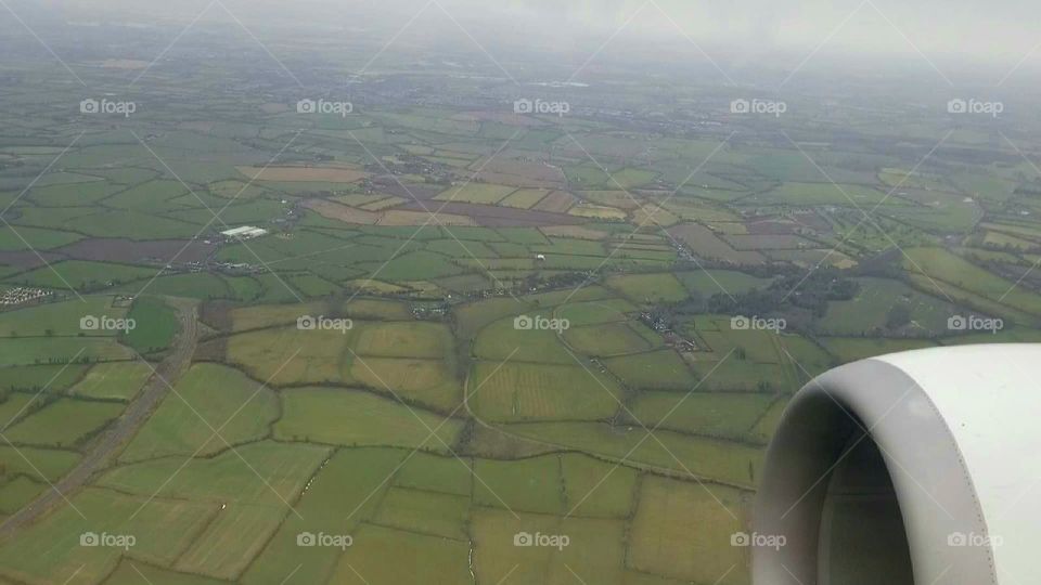 View from above, airplane window with jet engine on wing in view, looking out at shades of green colored farms and fields landscape, flying over Ireland.