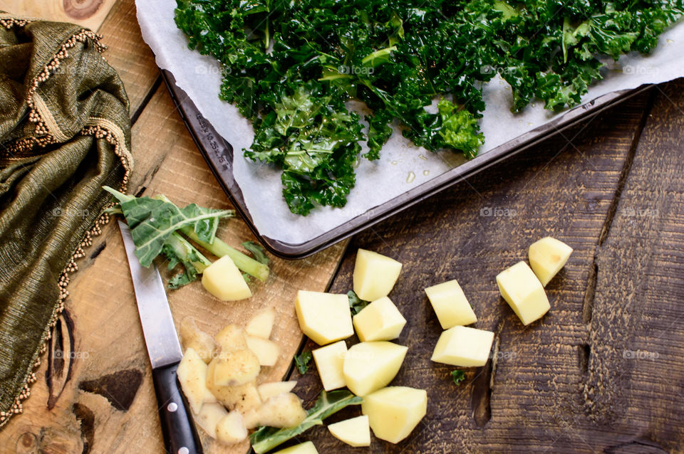 High angle view of healthy kale with olive oil on baking sheet on rustic wood cutting board with chopped potatoes preparing healthy autumn food crispy kale