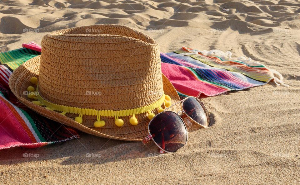 A straw hat, sunglasses and a Mexican beach coverlet lie on the sand by the sea on a clear sunny summer day, close-up side view.