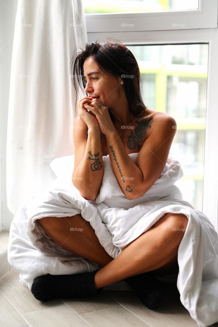 beautiful girl in tattoos, sitting by the window, wrapped in a white blanket. The girl looks out the window and thinks about life