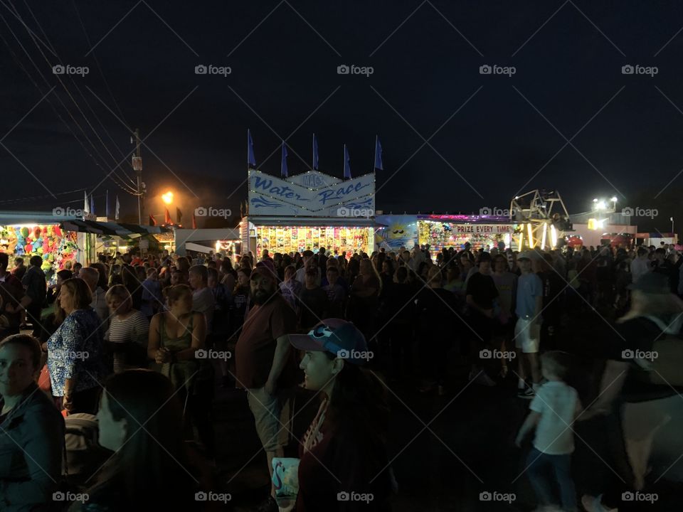 A crowd of people stand in front of carnival kiosks late at night with bright lights scattered around.