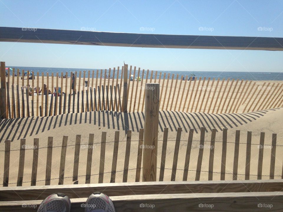 A photo of the beach from a bench on the Point Pleasant Beach boardwalk in NJ. The tips of my sneakers rest against the bottom of the boardwalk fence, the ocean and beach goers are in the distance, and the beach fence makes an interesting pattern. 