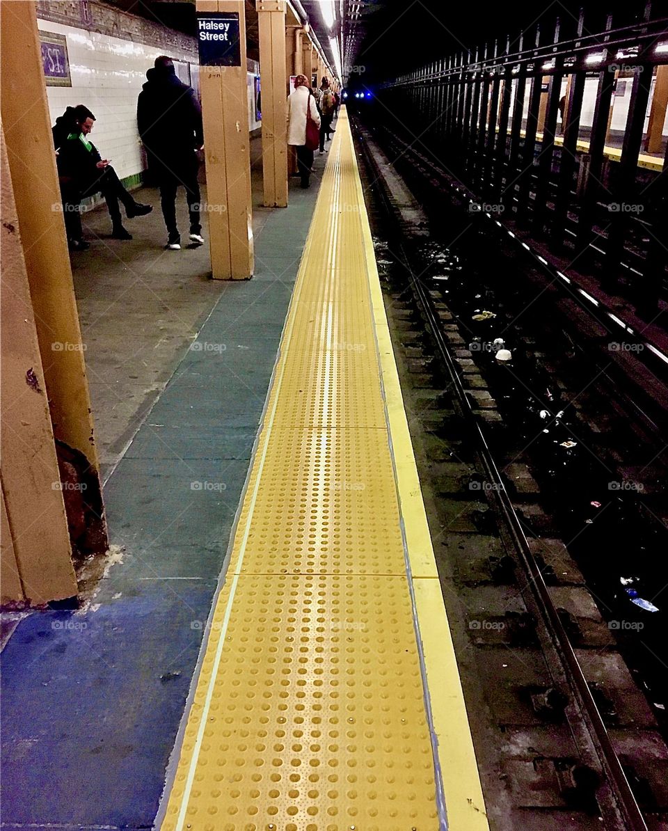 A subway station w a man waiting for the train to travel within NYC, the “Big Apple”. The bright yellow stripe at the edge of the platform indicates where it ends so no slip & fall accidents due to absent mindedness occur. 2021. Hypnotic Productions