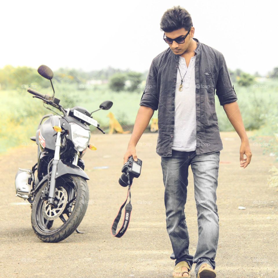 Photographer with motorbike.
View of young man walking and holding canon camera with motor bike parked in the background.