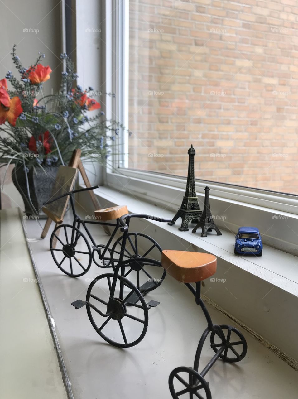 Still Life of poppies in a planter, two small Eiffel tower replicas, and bicycles on a window sill.