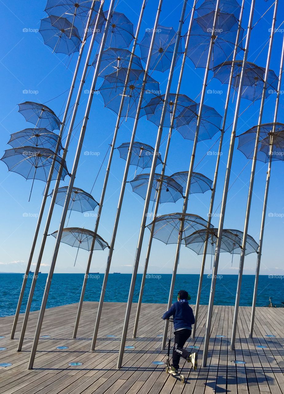 A boy with his scooter in front of thessaloniki's umbrellas.