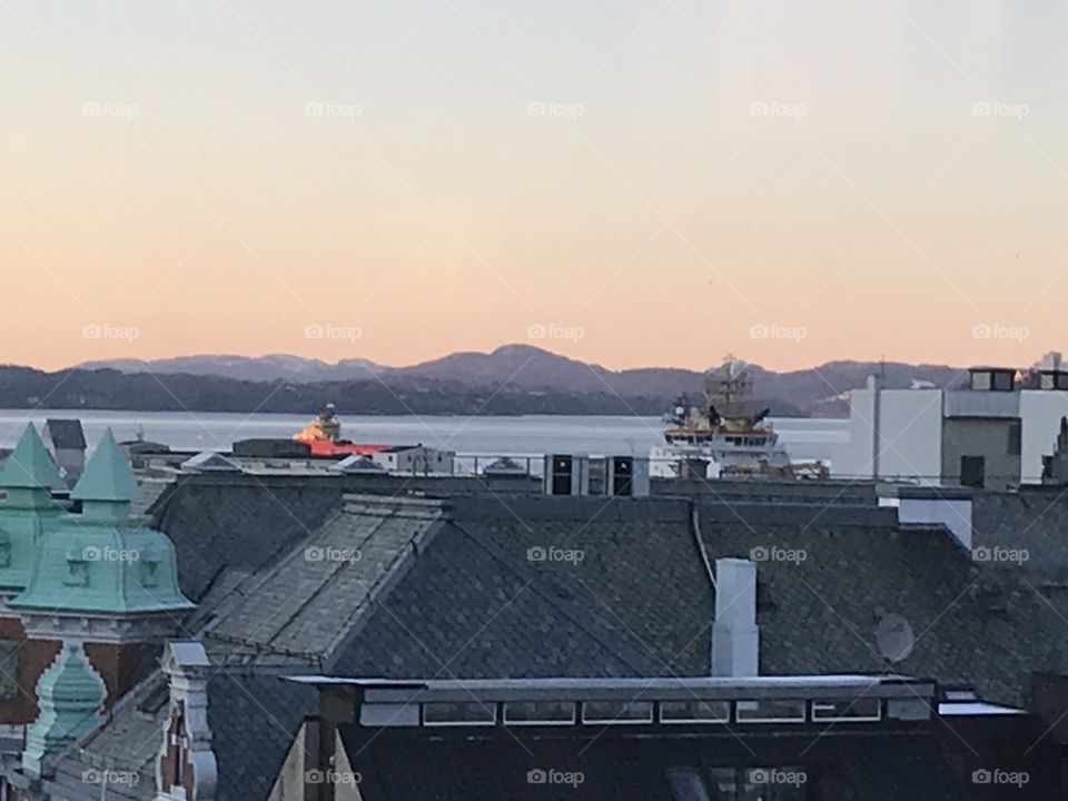 The sunset fades out over the mountains in the background. In front of the mountains we see some of the ships docked at that time. Before that some of the roofs in Bergen. Taken at a March evening in Bergen, Norway.