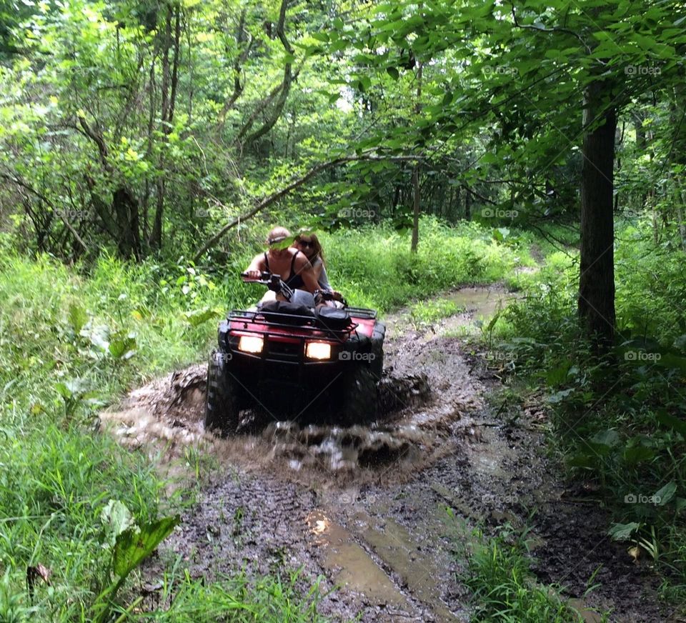 Two people riding a four wheeler on a very muddy path on a hot summer day in green forest. 