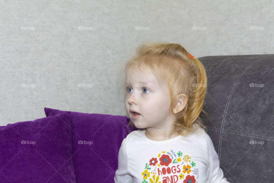 A small, red-haired girl is laughing at home on a gray sofa with purple pillows and a yellow background
