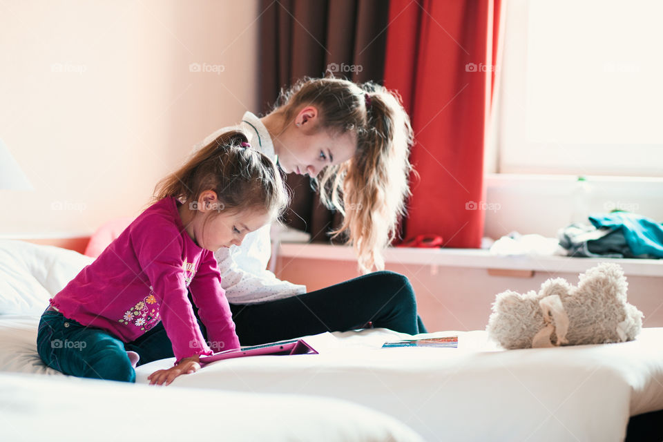 Teenage girl using mobile phone together with her little sister watching animated movie on tablet, both girls sitting in bed in bedroom