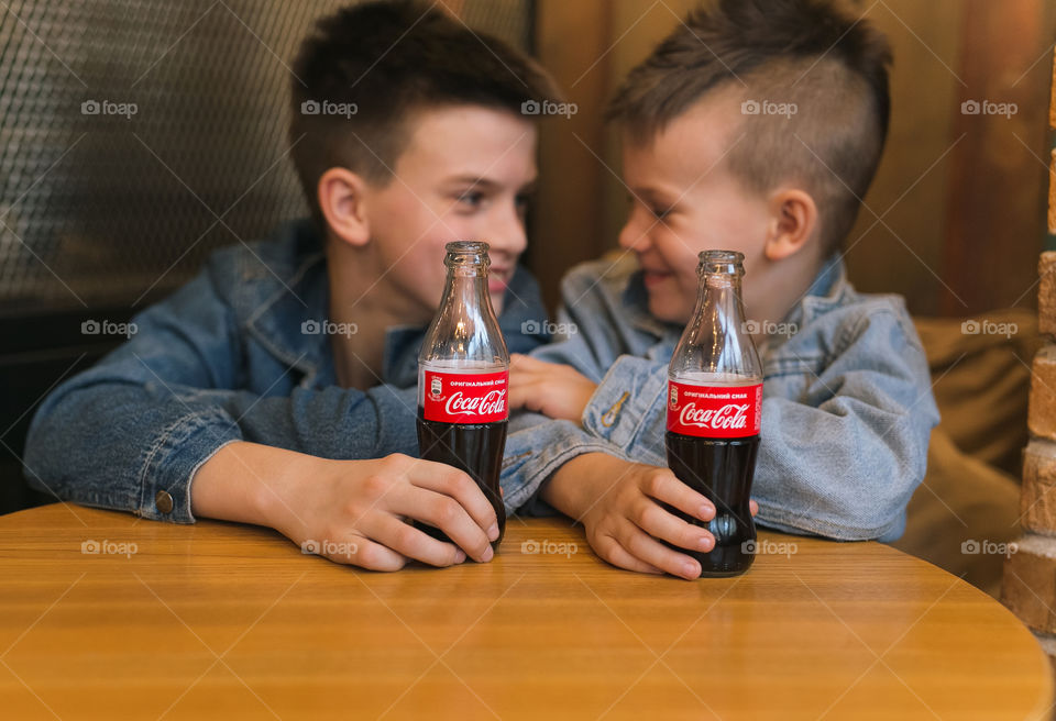 two lovely brothers, dressed in denim clothes, with beautiful hairstyles, are sitting in a cafe and drinking Coca-Cola.  communicate, laugh, smile.  two guys, two friends