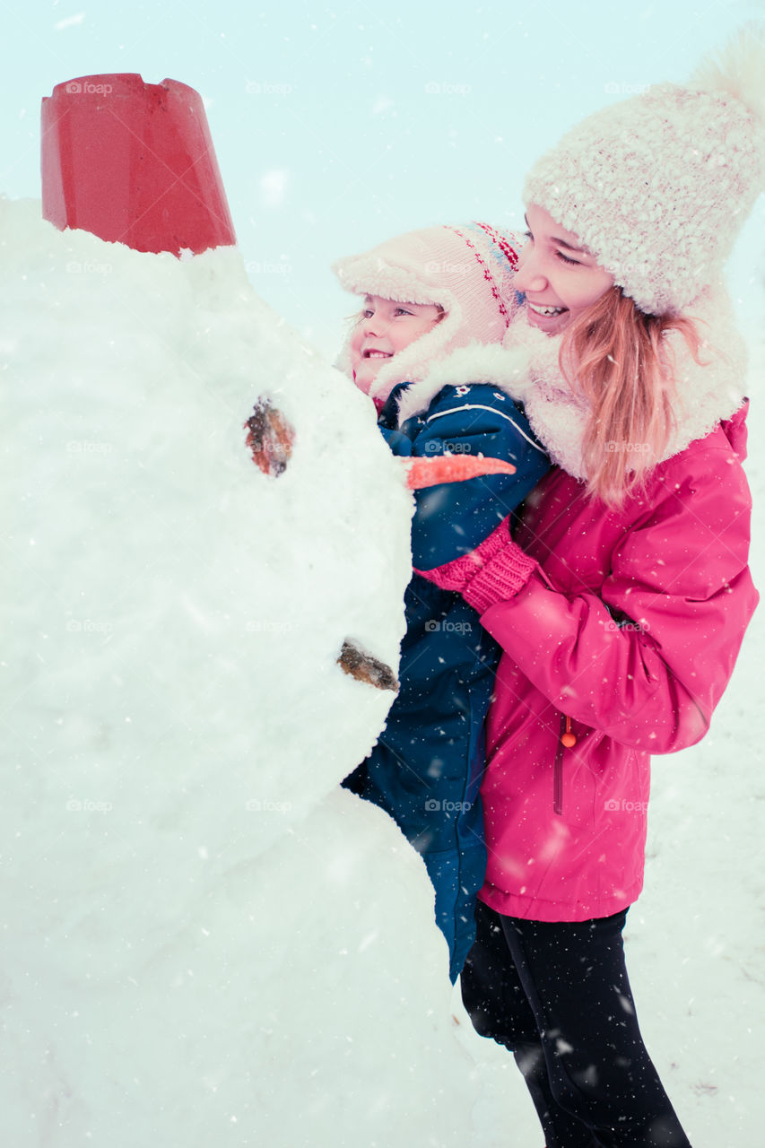 Girl and her little sister making a snowman, spending time together outdoors, having fun in wintertime