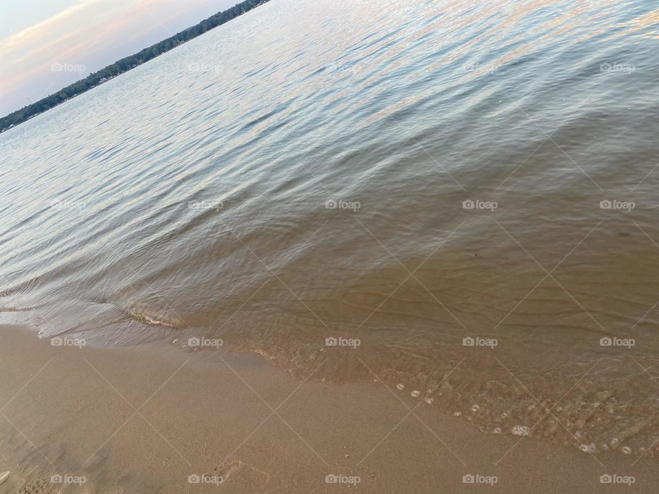 This image captures a close-up view of gentle waves lapping against a sandy beach. The water appears calm, and the shoreline is smooth with small bubbles forming at the edge where the water meets the sand, a peaceful and serene beach scene.