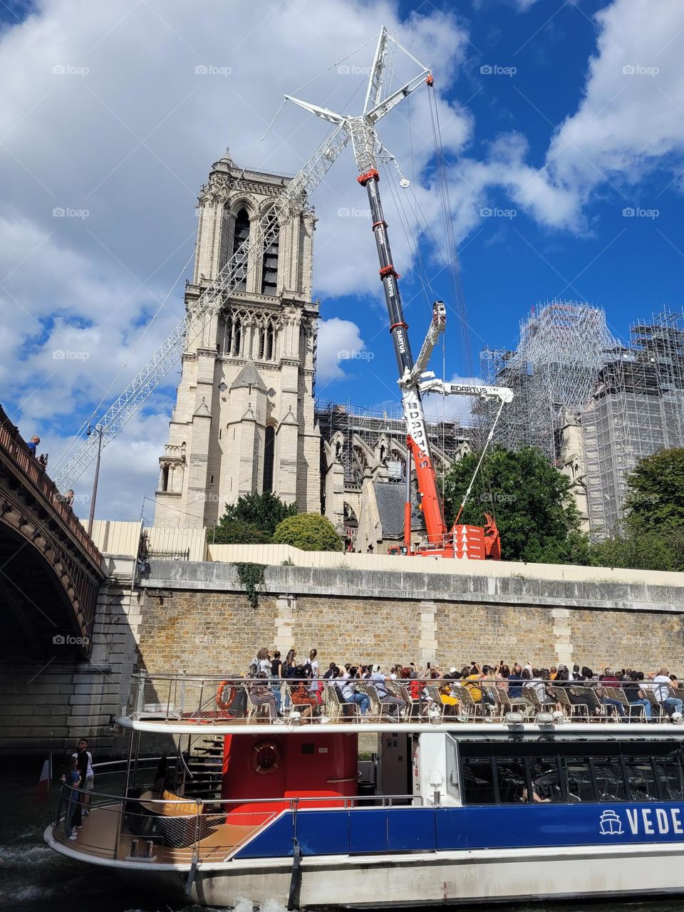 Boat full of tourists on Seine river watching Notre Dame church under reconstruction with complex crane and massive scaffolding.