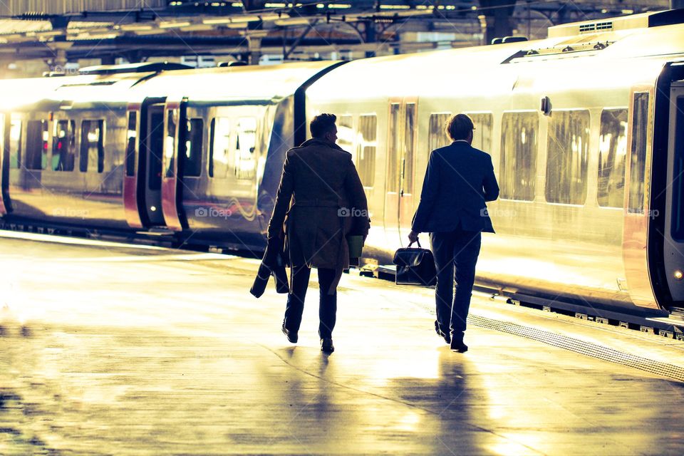 A pair of businessmen commuting at a railway station and walking along a sunlit platform before boarding a waiting train.