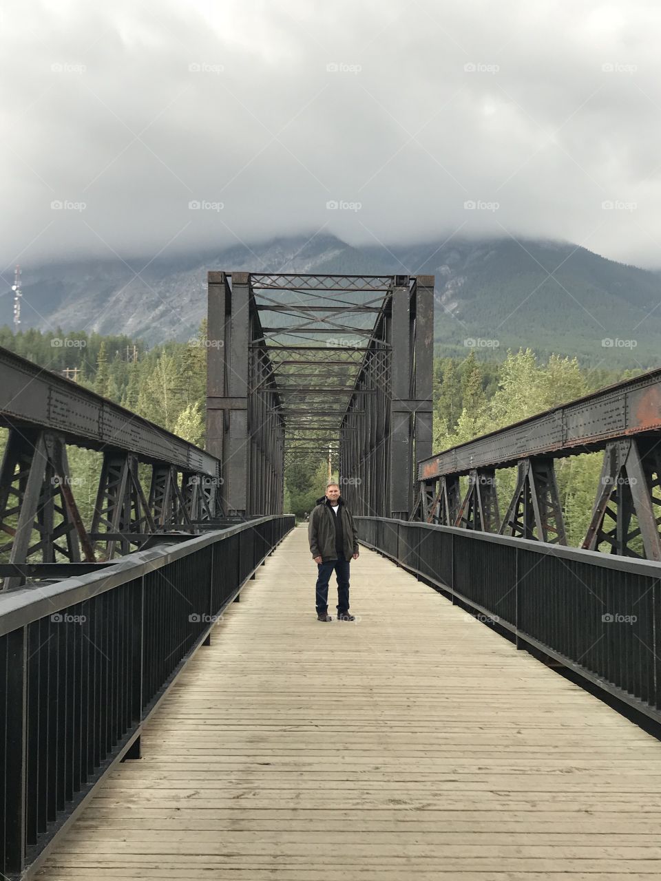 Man standing on a wooden and steel bridge in Canmore. Alberta, Canada.  The mountain range in the background has low rain clouds.
